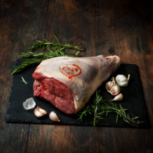 Raw, whole bone-in leg of lamb on a piece of slate with garlic and rosemary surrounding.