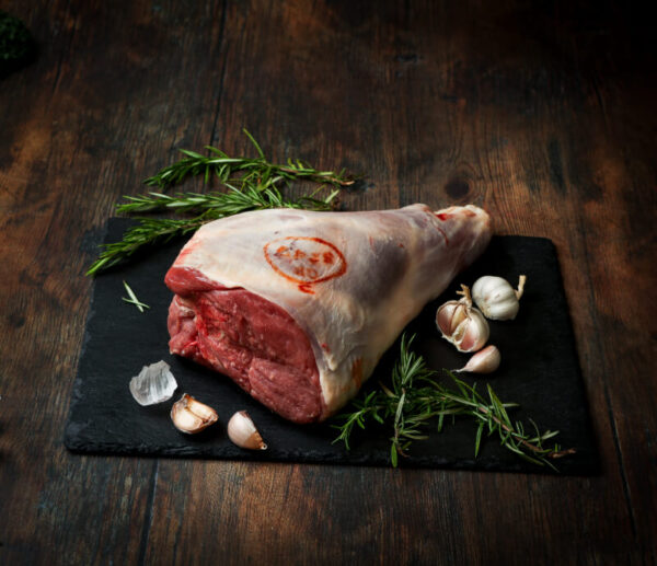 Raw, whole bone-in leg of lamb on a piece of slate with garlic and rosemary surrounding.